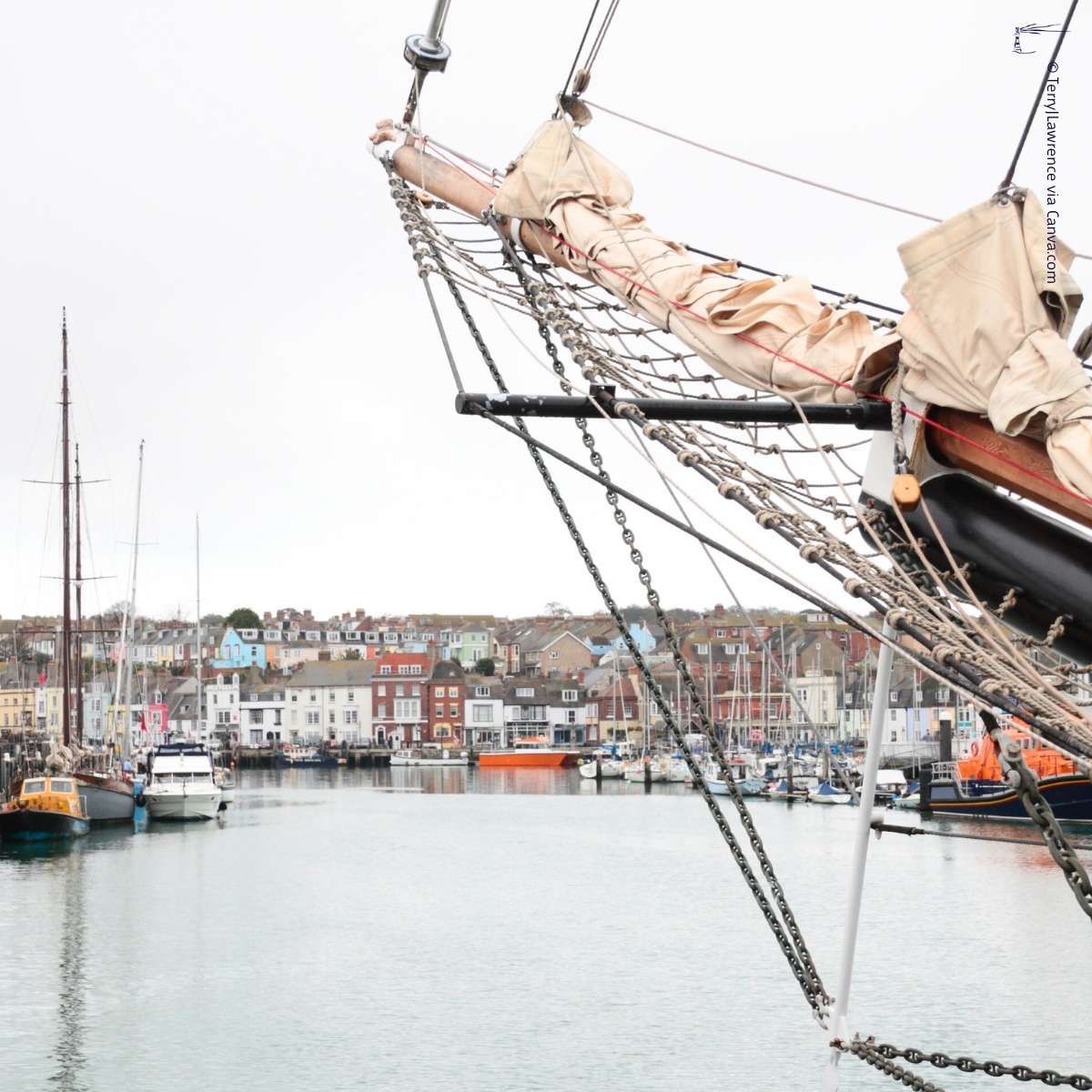 Im Vordergrund ein angedocktes Segelboot mit Bug und Takelage, im Hintergrund ein Hafen mit verschiedenen Booten und eine Stadt mit bunten Gebäuden.